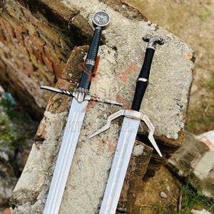 May include: Two silver-toned metal swords with black grips. The left sword features a circular pommel with a wolf emblem and a straight crossguard. The right sword has a stylized animal head pommel and a unique, angular crossguard design.
