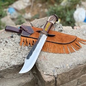 Peut inclure: Un grand couteau de chasse avec une lame en acier poli et un manche en corne et en cuir. Le couteau est accompagné d'un étui en cuir marron avec des franges et une boucle de ceinture.