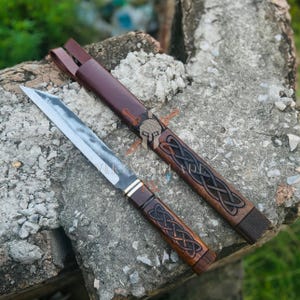 May include: A handcrafted knife with a wooden handle and a stainless steel blade, alongside a matching leather sheath. The handle and sheath feature an intricate Celtic knot design. The knife is displayed on a rough stone surface.