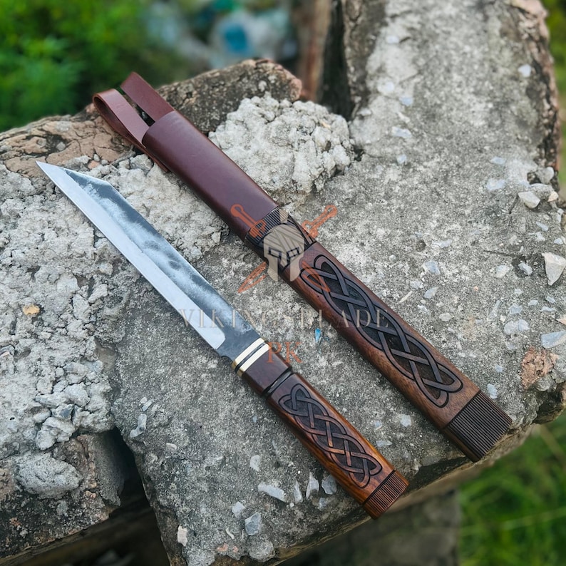 May include: A handcrafted knife with a wooden handle and a stainless steel blade, alongside a matching leather sheath. The handle and sheath feature an intricate Celtic knot design. The knife is displayed on a rough stone surface.