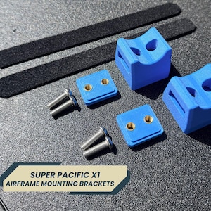 Può includere: Un assortimento di staffe di montaggio per la cellula e hardware. L'immagine mostra due staffe blu a forma di cubo, due staffe quadrate blu più piccole, due strisce nere e diverse viti argentate. Il testo "SUPER PACIFIC X1 AIRFRAME MOUNTING BRACKETS" è visibile.
