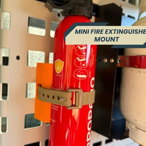 May include: A red mini fire extinguisher is held in place by a tan strap and mounted on a white metal grid. The text "MINI FIRE EXTINGUISHER MOUNT" is displayed on a banner. An orange card is also attached.