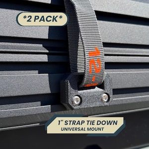 May include: Close-up of a black strap tie-down with a universal mount. The strap is grey with orange text and is attached to a black surface. The text "*2 PACK*" is in a cream-coloured oval. The text "1" STRAP TIE DOWN UNIVERSAL MOUNT" is in a blue and cream-coloured rectangle.