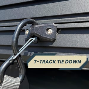 May include: Close-up of a black T-track tie-down system. The system includes a black metal carabiner attached to a black plastic component secured with screws. The text "T-TRACK TIE DOWN" is displayed on a banner.