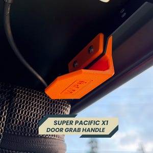 Puede incluir: Manija de puerta de color naranja brillante con el texto "WPU" y "SUPER PACIFIC X1 DOOR GRAB HANDLE" impreso. La manija está montada con dos tornillos. El producto está diseñado para ser instalado en un vehículo.