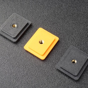 May include: Three small, square, layered objects with a central hole. Two are dark gray, and one is bright yellow. Each has a metallic insert in the center. The objects are arranged on a textured, dark surface.