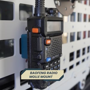May include: Black Baofeng radio mounted on a white panel with a blue bracket. The radio has an orange button and a small screen. The text "BAOFENG RADIO MOLLE MOUNT" is displayed on a banner below the radio.