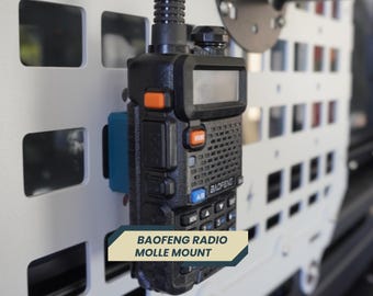 May include: Black Baofeng radio mounted on a white panel with a blue bracket. The radio has an orange button and a small screen. The text "BAOFENG RADIO MOLLE MOUNT" is displayed on a banner below the radio.