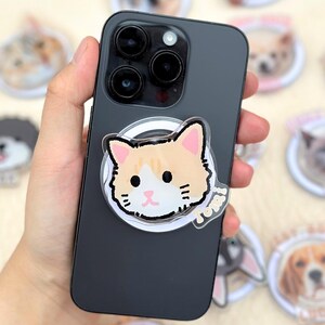 Puede incluir: Un teléfono inteligente negro con un agarre para teléfono con temática de gato adjunto. El agarre presenta un gato de dibujos animados con pelaje naranja y blanco. Otros agarres para teléfono con temática de mascotas con caras de perros y gatos están dispersos.