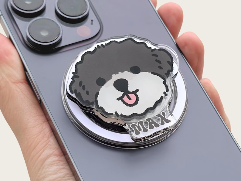 May include: A silver phone ring holder with a clear plastic overlay featuring a cartoon black and white dog with the name "MAX" printed on the bottom.