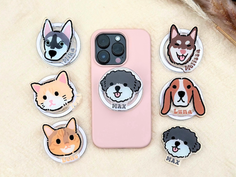 May include: A pink smartphone case with a variety of phone grips featuring different dog and cat illustrations. The grips are clear with white outlines and each has a name printed on the bottom. The names are Mika, Luna, Max, and Eiko.