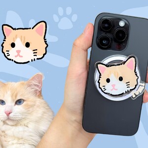 May include: A black iPhone case with a clear phone ring holder featuring a cartoon orange and white cat face. The phone ring holder has the word "Meow" printed on it.