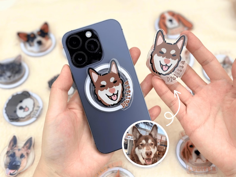 May include: A close-up of a phone case with a clear phone grip featuring a cartoon dog with the name "Melissa" printed on it. The phone case is dark blue and has a camera lens on the back. The phone is being held by a person's hand. There are other phone grips with cartoon dogs on them in the background.