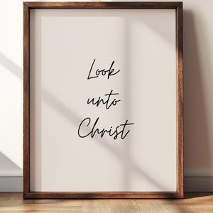 May include: A framed print with the words "Look unto Christ" in black script. The frame is a dark brown color, and the background is a light beige. The print is displayed on a wooden floor.