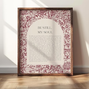 May include: A framed print with the text "BE STILL, MY SOUL" in a decorative font. The print has a cream-colored background with a floral border and text in a deep red color. The frame is a warm brown color.