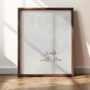 May include: A rectangular wall art print encased in a dark brown wooden frame. The print features a light gray, watercolor-style background with the dark brown cursive text "Walk with Me" centered in the lower portion of the composition.