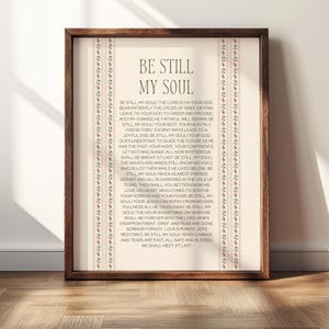 May include: A framed wall art print featuring the hymn Be Still My Soul in black serif text on a cream background. The design includes two vertical decorative borders with a repeating floral pattern in red and green within a dark wood frame.