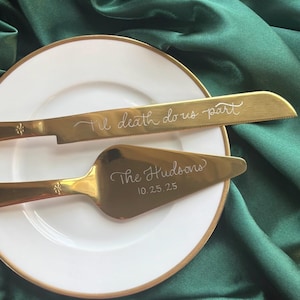 May include: A gold-colored cake knife and server set on a white plate with a gold rim. The knife is engraved with "'Til death do us part," and the server with "The Hudsons" and the date 10.25.25. The background is a dark green satin fabric.