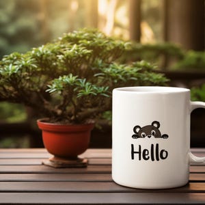 May include: White ceramic mug with a black bear graphic peeking over the top and the word "Hello" in black. The mug is on a wooden table with a bonsai tree in the background.