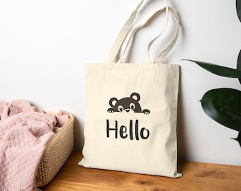 May include: Cream-colored tote bag with a black bear graphic and the word "Hello" in a bold, black font. The bag has two handles and is made of a sturdy fabric. A pink blanket is in a basket to the left of the bag.