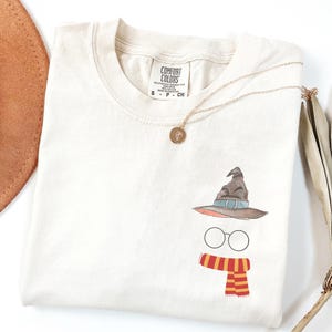 May include: Cream-colored t-shirt with a watercolor illustration of a wizard hat, glasses, and a striped scarf. A gold necklace with a pendant rests on the shirt. The shirt has a Comfort Colors tag.