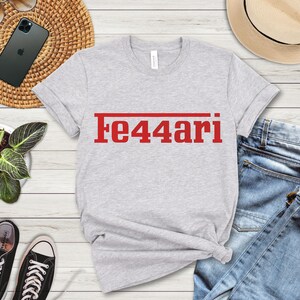 May include: A light gray t-shirt with the red text "Fe44ari" printed across the chest. The shirt is made of a soft material and has a crew neck. The shirt is paired with blue jeans and black sneakers.