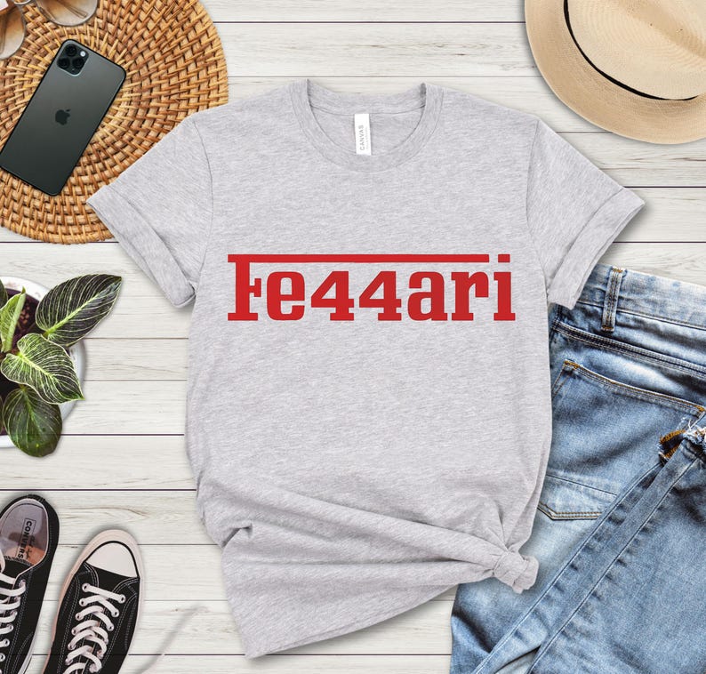 May include: A light gray t-shirt with the red text "Fe44ari" printed across the chest. The shirt is made of a soft material and has a crew neck. The shirt is paired with blue jeans and black sneakers.