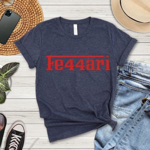 May include: Navy blue t-shirt with the red text "Fe44ari" printed across the chest. The shirt is made of a soft material and has a classic crew neck design. The shirt is folded on a light wood surface.