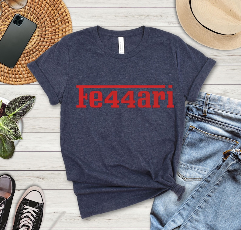 May include: Navy blue t-shirt with the red text "Fe44ari" printed across the chest. The shirt is made of a soft material and has a classic crew neck design. The shirt is folded on a light wood surface.