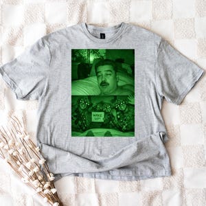 May include: A gray t-shirt featuring a green-tinted image. The image shows a man in bed and soldiers holding a sign that says "WAKE UP!". The shirt is made of a soft material.