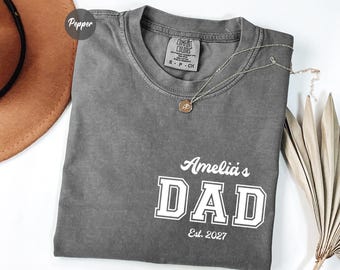 May include: A charcoal gray short-sleeve t-shirt with a crew neckline. The shirt features white screen-printed text that reads Amelia's DAD Est. 2027 in a combination of script and block lettering. The garment has a Comfort Colors brand label.