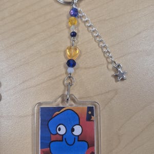 May include: A clear acrylic keychain featuring a blue cartoon character with a smiling face. The keychain has a silver keyring, a chain, and a star charm. Decorative beads in yellow, blue, and clear are strung along the chain.