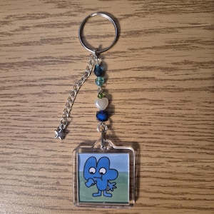 Puede incluir: Un llavero acrílico transparente con un personaje de dibujos animados azul con el número cuatro. El llavero tiene una cadena plateada con un dije de estrella y está adornado con cuentas azules, verdes y blancas. El personaje está sobre un fondo de cielo azul y hierba verde.