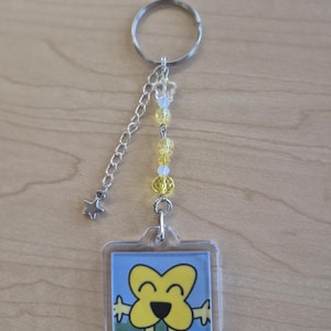 Puede incluir: Un llavero acrílico transparente con un colgante cuadrado que presenta un personaje de dibujos animados amarillo con una nariz y extremidades negras. El llavero tiene una cadena plateada, un dije de estrella y cuentas amarillas y transparentes.