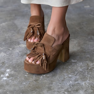 Pode incluir: Sandálias plataforma de camurça castanha com salto bloco e detalhes de franjas. As mulas de bico aberto têm um laço e uma sola de plataforma grossa. Os sapatos são de cor castanha quente e têm um estilo vintage e retro.