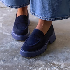 May include: Navy blue suede loafers with a chunky, lavender-colored sole. The shoes have a classic penny loafer design with a rounded toe and a strap across the vamp. The shoes are worn with blue denim pants.