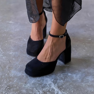 May include: Black platform heels with a square toe and ankle strap. The heels have a thick block heel and a suede-like texture. The shoes are worn with a black garment.