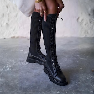 May include: Black knee-high boots featuring a ribbed knit shaft and a lace-up design. The boots have a leather toe cap and a thick, flat sole. A person is adjusting the boots, wearing gold rings and a bracelet.