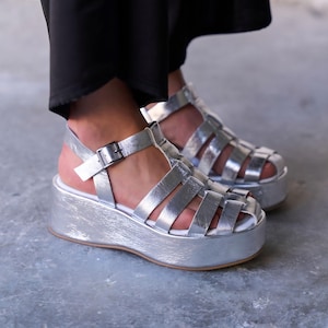 May include: Silver platform sandals with a cage design. These sandals feature a thick sole and a buckle fastening on the ankle strap. The metallic finish gives a modern look to these shoes.