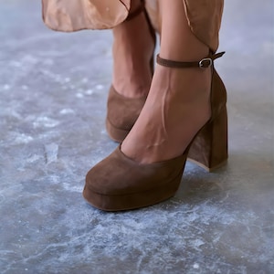 May include: Brown suede platform heels with a rounded toe and ankle strap. The block heel provides stability, and the platform sole adds height. These shoes are a stylish choice for various occasions.