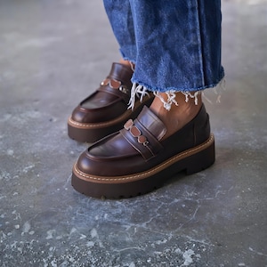 May include: Brown leather platform loafers with decorative straps. The shoes have a chunky sole and a classic penny loafer design. The image shows the shoes from a low angle, highlighting their style and construction. The shoes are paired with blue denim jeans.