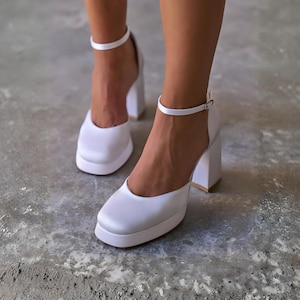 Könnte beinhalten: Weiße Satin-Plateau-Heels mit eckiger Zehenpartie und Knöchelriemen. Die Blockabsätze haben einen goldenen Besatz. Die Schuhe sind mit geschlossener Zehenpartie und dicker Plateausohle ausgestattet.