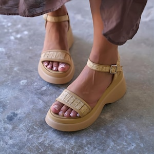 May include: Tan platform sandals with woven raffia straps and ankle straps. The sandals have a rounded toe and a buckle closure. The platform sole is a solid tan color. The sandals are a neutral color and suitable for various outfits.