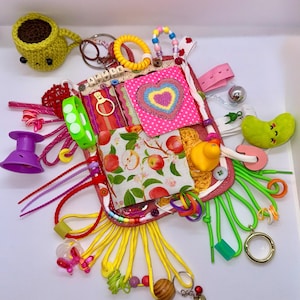 May include: A sensory activity board with various textures and elements. Includes a crocheted yellow coffee cup, a pink heart-shaped patch, a green bracelet, a yellow rubber duck, and multiple colorful strings and beads. The board is designed for tactile exploration.