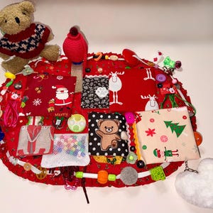 Puede incluir: Un juguete sensorial rojo hecho a mano con varias texturas y elementos interactivos. Presenta un oso de peluche, cuadrados de tela con temática navideña con Papá Noel y renos, botones y un dije suave en forma de corazón blanco. Diseñado para la exploración táctil.