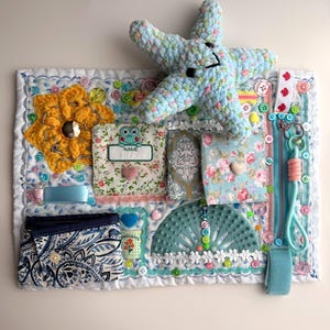 May include: A sensory board with various textures and interactive elements. Features a light blue crocheted starfish, a yellow flower, buttons, zippers, and patterned fabrics. Includes a small label with the word "NAME".