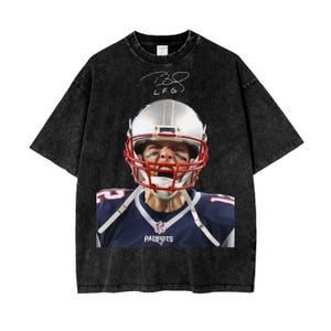 Peut inclure: T-shirt noir à l'aspect délavé avec une photo de Tom Brady en uniforme de football, portant un casque et regardant sur le côté. Le texte "LF G" est écrit en blanc au-dessus de la photo.