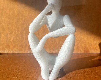 May include: A white, abstract sculpture of a person sitting with their chin resting on their hand. The sculpture is made of a smooth, textured material.