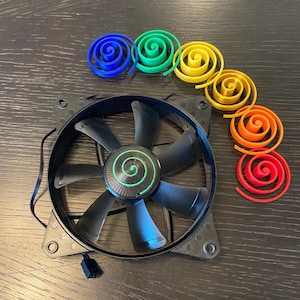 May include: A black computer cooling fan with seven blades and a green spiral emblem on the center hub, accompanied by six separate spiral-shaped plastic components in blue, green, yellow, gold, orange, and red arranged in a curved line.