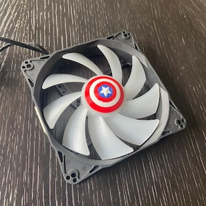 May include: A square computer cooling fan featuring a dark gray frame and nine translucent white blades. The center hub is decorated with a circular red, white, and blue shield emblem featuring a prominent white star in the middle.